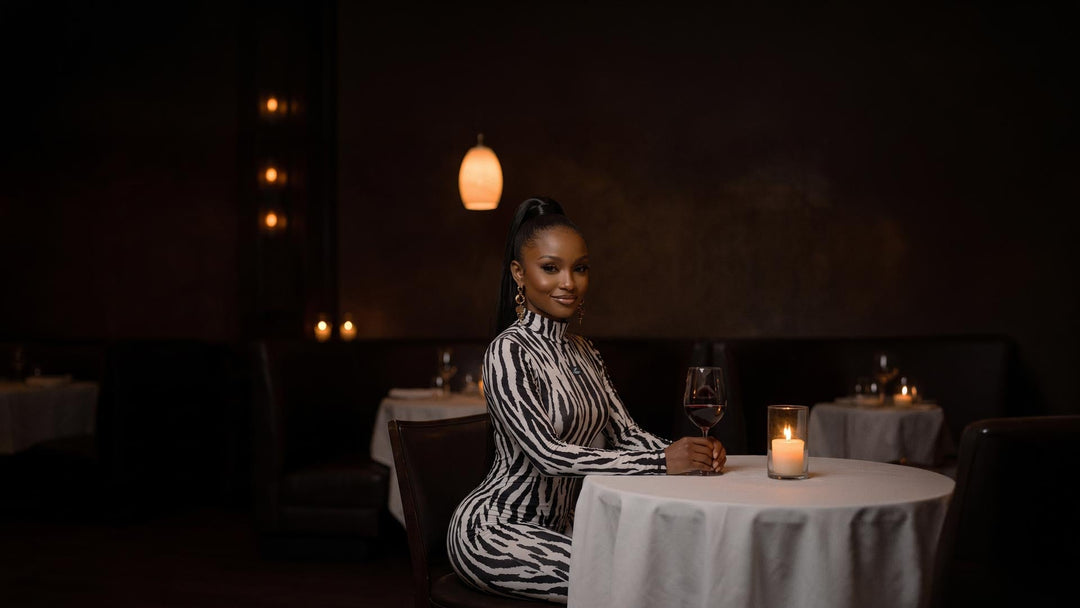 Woman in a black and white zebra print long sleeve jumpsuit seated at a candlelit restaurant table with a glass of red wine