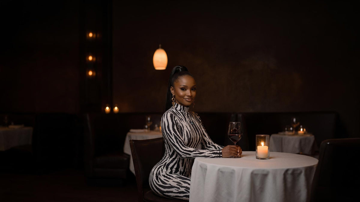 Woman in a black and white zebra print long sleeve jumpsuit seated at a candlelit restaurant table with a glass of red wine