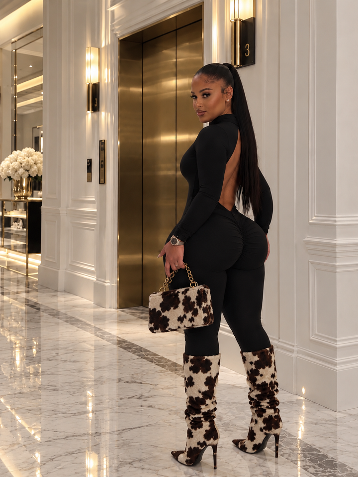 Back view of the Dark Desire Jumpsuit showing the open back scooped to the waist and the ruched center seat, worn with black and cream print knee boots