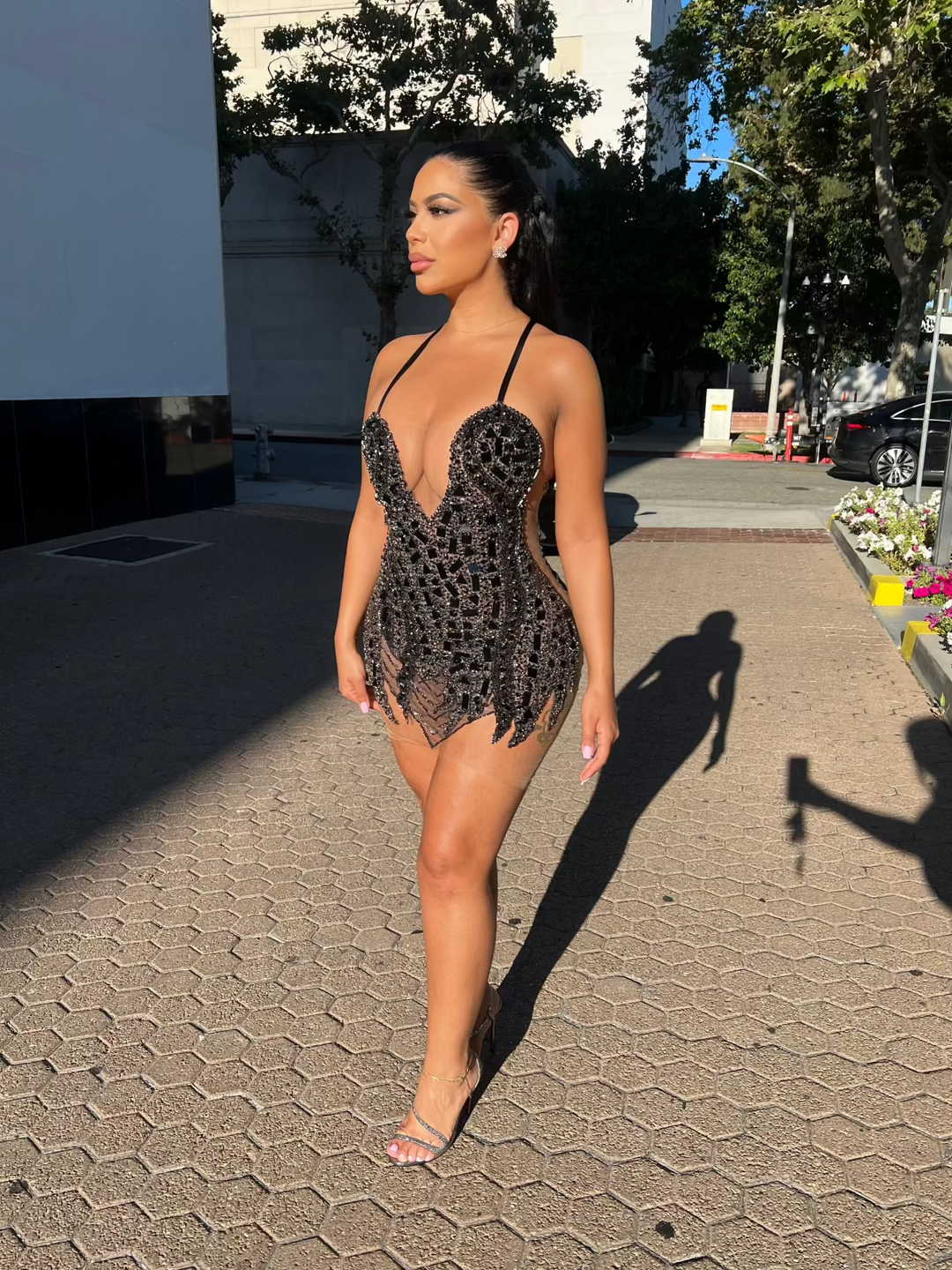 A woman wearing a black sleeveless mini dress with hand sewn diamonds and mirrors on the front, standing on a sidewalk.