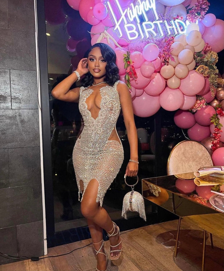 A person wearing a sleeveless, sequined dress with a deep neckline and embellished with sparkling stones, standing in front of a 'Happy Birthday' balloon setup.